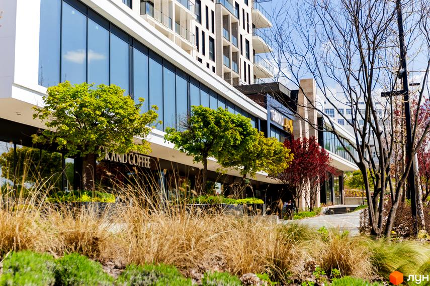 Фасад Житлового комплексу Grand Hills з великими скляними вікнами та білими балконами. Перед будинком розташовані зелені дерева, доглянуті газони та сухі трави.