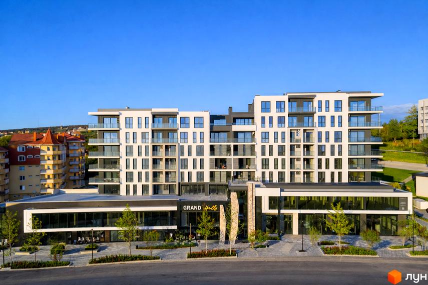 Житловий комплекс Grand Hills з біло-сірим фасадом, балконами та великими вікнами. Перед будинком облаштована територія з деревами та пішохідними доріжками.