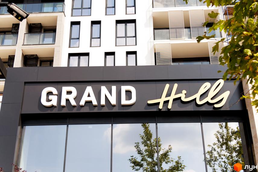 Фасад Житлового комплексу Grand Hills з білими стінами, темними вікнами та балконами. На вивісці назва проєкту, поруч зелені дерева.