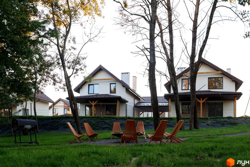 Будинки Котеджного містечка Буде Дім. Деревач мають світлі фасади, коричневі дахи та дерев'яні опори. На передньому плані зелений газон з дерев'яними стільцями та мангалом.