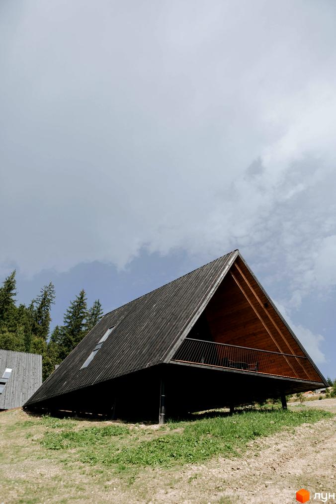 Комплекс відпочинку УЛІС Ворохта. Дерев'яний будинок трикутної форми з балконом на палях, оточений зеленою травою та деревами.