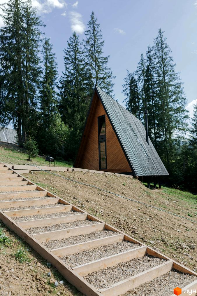 Дерев'яний будинок А-подібної форми з темним металевим дахом та високим вікном. До нього ведуть дерев'яні сходи, а навколо ростуть хвойні дерева.