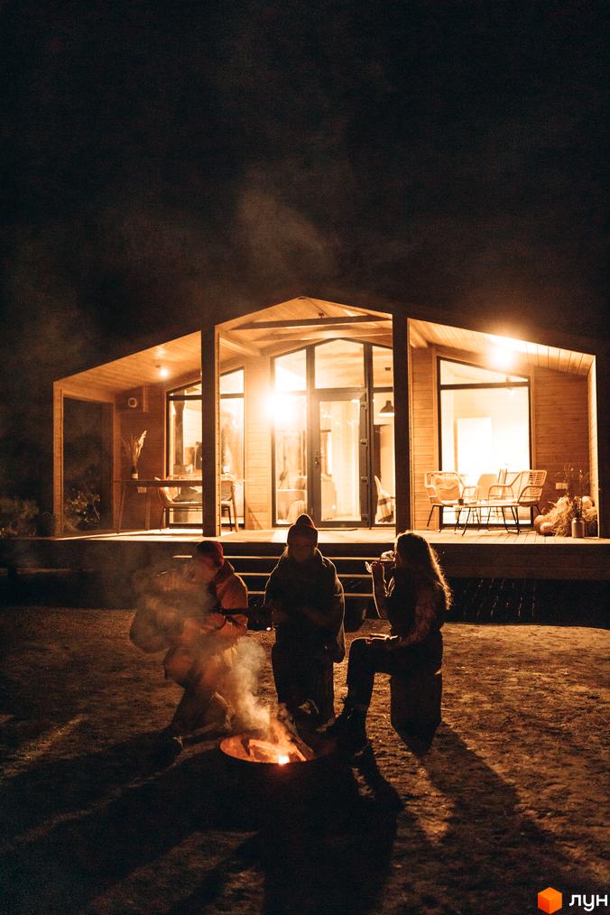 Комплекс відпочинку УЛІС Віняви з дерев'яним фасадом та великими вікнами вночі, люди біля багаття на прибудинковій території.