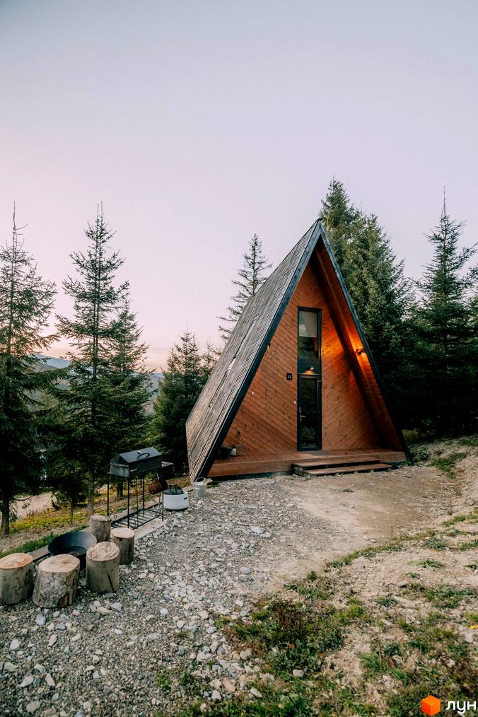 Будинок комплексу відпочинку УЛІС Ворохта у формі трикутника, оточений ялинами. Біля будинку розташовані дрова, мангал та місце для відпочинку.