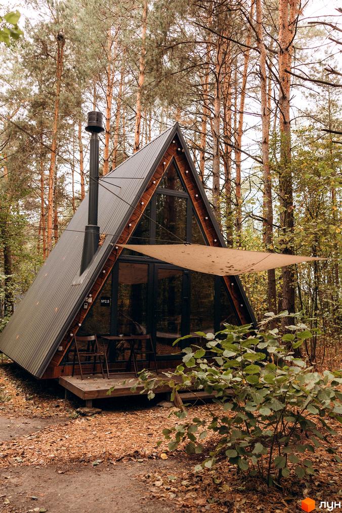 Комплекс відпочинку УЛІС Дудки: трикутний будинок з темним дахом, великими вікнами та дерев'яною терасою. Розташований у сосновому лісі.