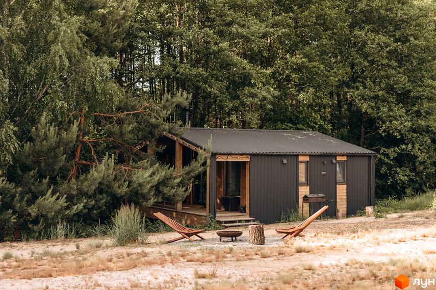 Темний будинок з дерев'яними елементами та чорним дахом. Перед ним розташовані дерев'яні шезлонги, вогнище та ліс на задньому плані.