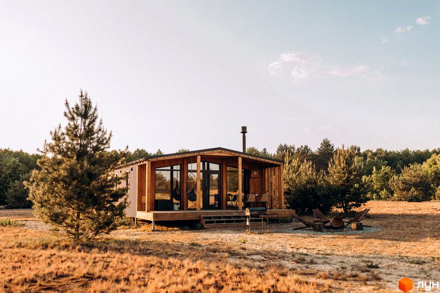 Дерев'яний будинок з панорамними вікнами та терасою серед соснового лісу. Поруч з будинком облаштована зона відпочинку з вогнищем.