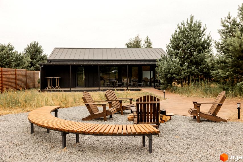 Комплекс відпочинку УЛІС Дудки з темною будівлею та дерев'яними меблями на території. Навколо будівлі розташовані зелені дерева та галькове покриття.