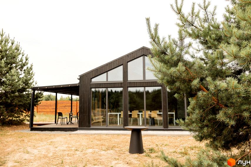 Темний будинок з панорамними вікнами та гострим дахом Комплекс відпочинку УЛІС Дудки. Тераса з меблями та сосни на піщаній ділянці.