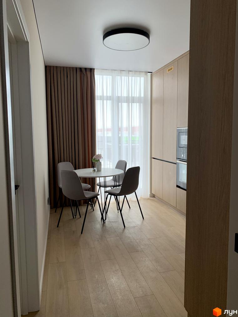 Інтер'єр кухні-їдальні в Житловому комплексі Fine Flats: дерев'яні меблі, вбудована техніка, обідній стіл та стільці. Світла підлога, вікно з шторами.