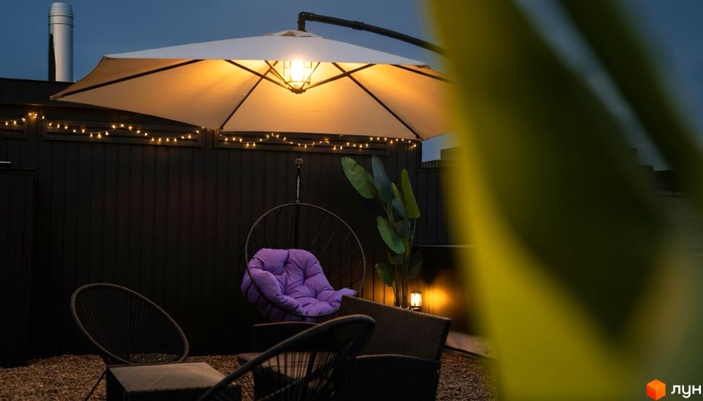 Затишна тераса з підвісним кріслом та вуличними меблями під навісом. Територія ЖК Долішній освітлена гірляндами.