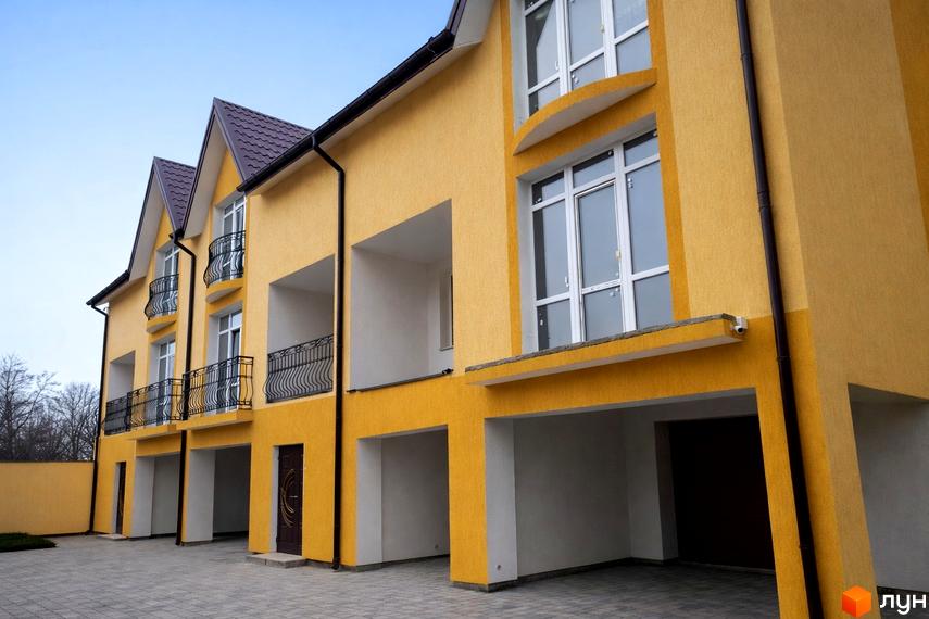 Зовнішній вигляд жовтого житлового комплексу з балконами та гаражами. Прибудинкова територія вимощена плиткою.