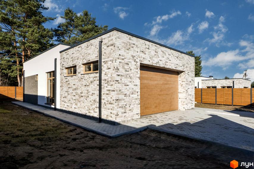 Котеджне містечко LOTUS. Зовнішній вигляд будинку з цегляним фасадом, дерев'яними вікнами та гаражними воротами. Навколо будинку вимощена бруківкою прибудинкова територія та зелена зона.