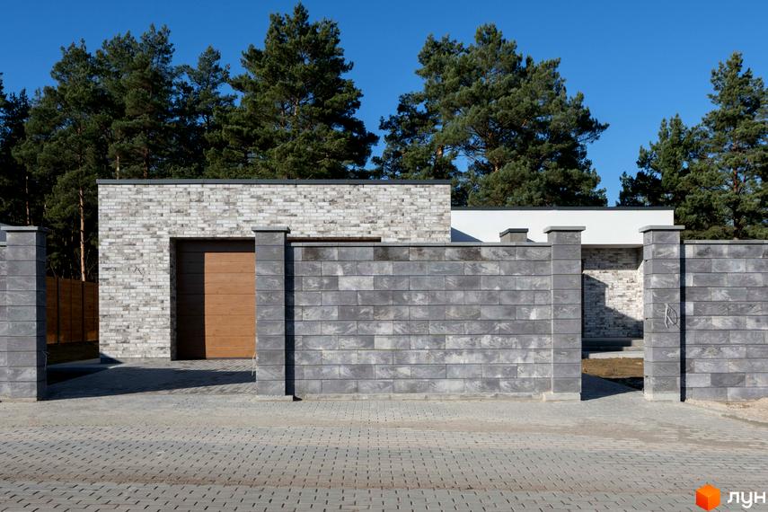 Котеджне містечко KM LOTUS: фасад будинку з цегли та дерев'яні ворота, огороджений парканом з сірого каменю. Прибудинкова територія вимощена бруківкою.