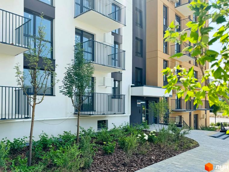 Новобудова ЖК Садова Алея з білими та бежевими фасадами, балконами з чорними огорожами та доглянутою прибудинковою територією з деревами та кущами.