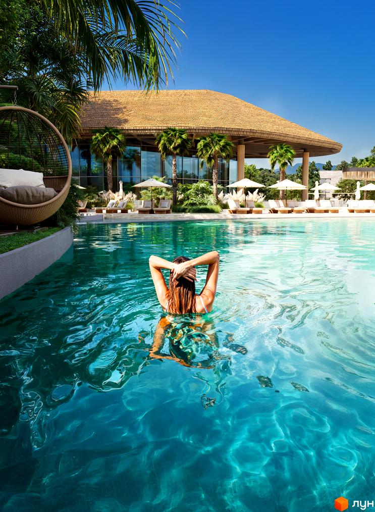 Апарт-готель ONOKY River Club з великим будинком під солом'яним дахом та панорамними вікнами. Перед ним басейн, лаунж-зона та пальми.