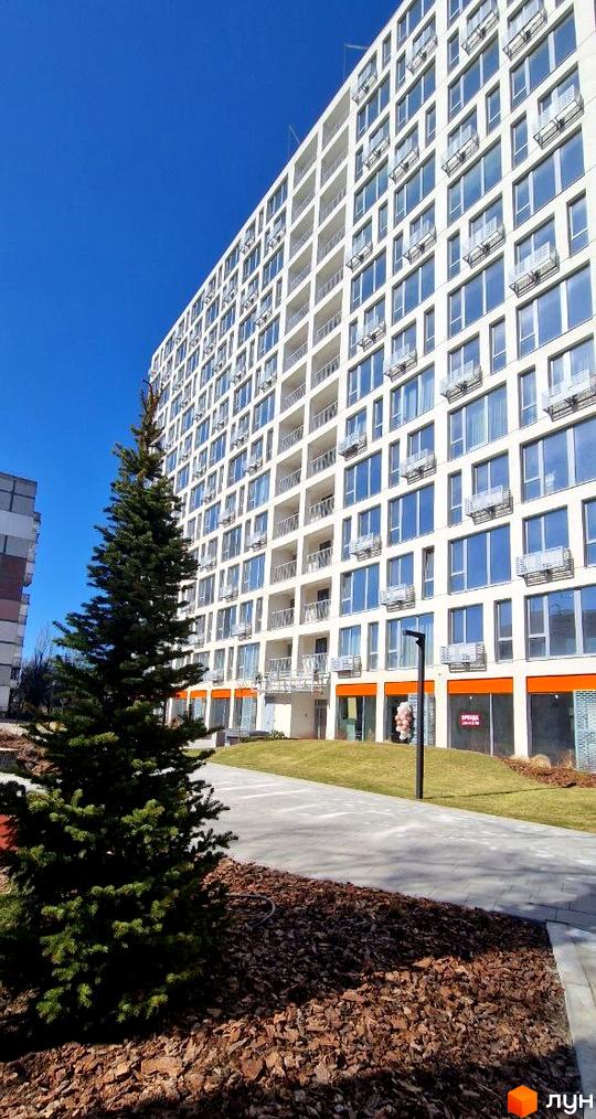 Багатоповерховий білий будинок Житлового комплексу Вертикаль з великими вікнами та балконами. Прибудинкова територія має доріжку, зелений газон та ялинку.