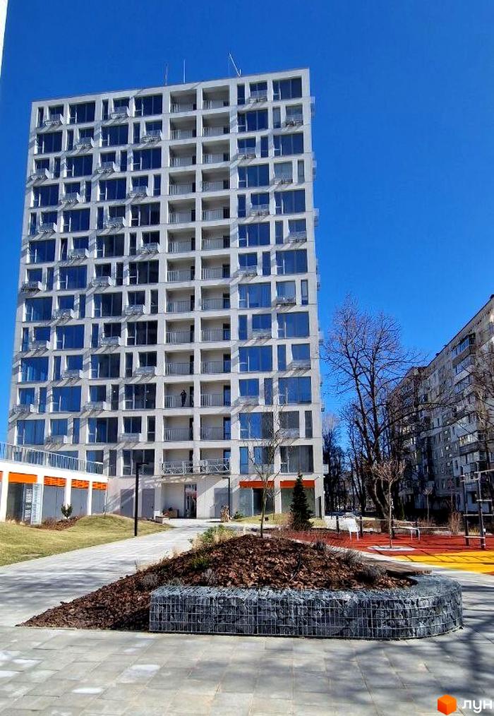 Житловий комплекс Вертикаль – висотна новобудова з численними балконами та панорамними вікнами на тлі блакитного неба. Поруч розташована прибудинкова територія з доглянутим газоном та декоративною клумбою.