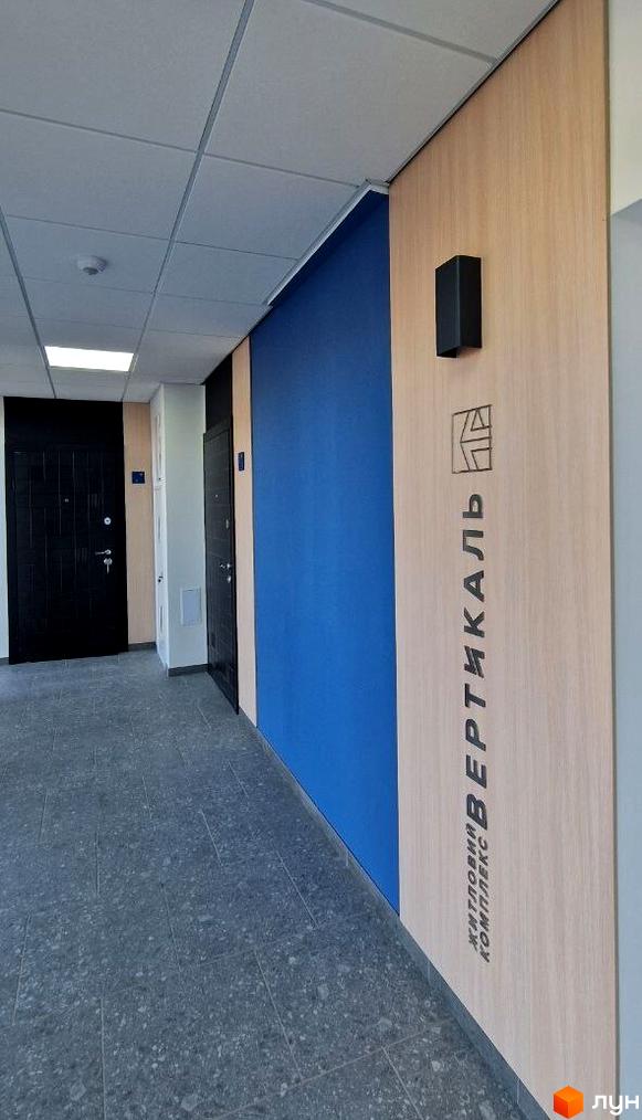 Коридор новобудови Житлового комплексу Вертикаль: стіни з дерев'яними та синіми панелями, темні двері, сіра плитка на підлозі.