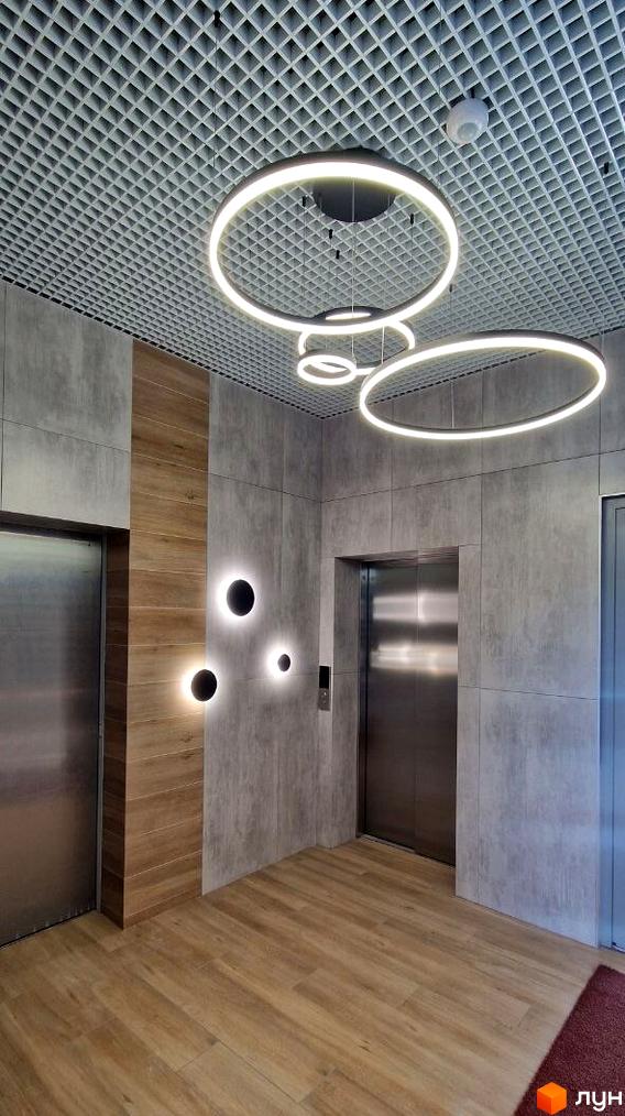 Житловий комплекс Вертикаль: інтер'єр ліфтового холу з сірими стіновими панелями, дерев'яними акцентами, круглими світильниками та двома ліфтами.