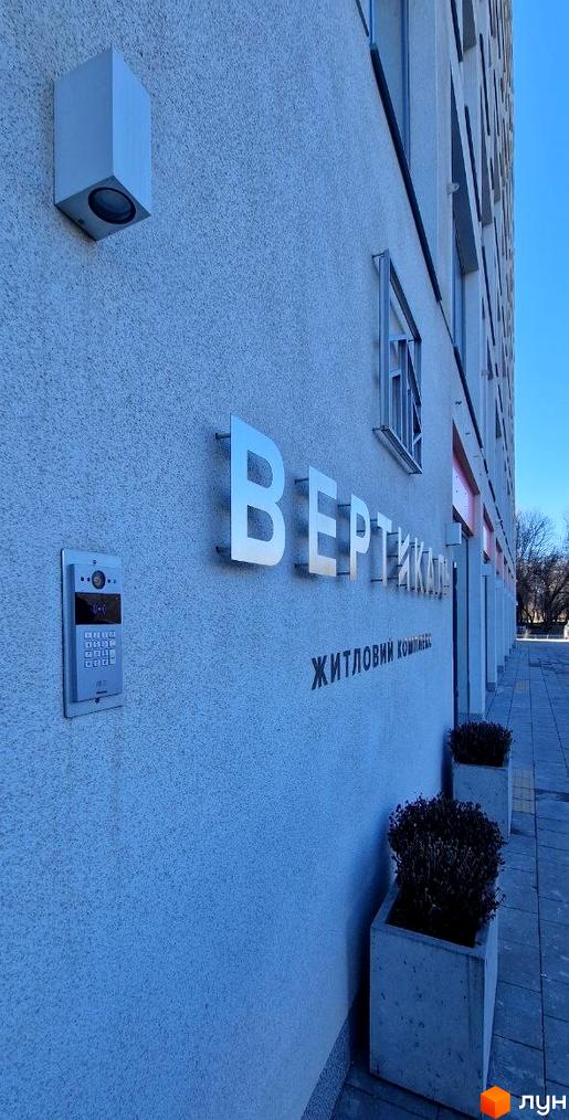 Зовнішній вигляд новобудови ЖК Вертикаль з написом на стіні та прибудинковою територією з рослинами в горщиках.