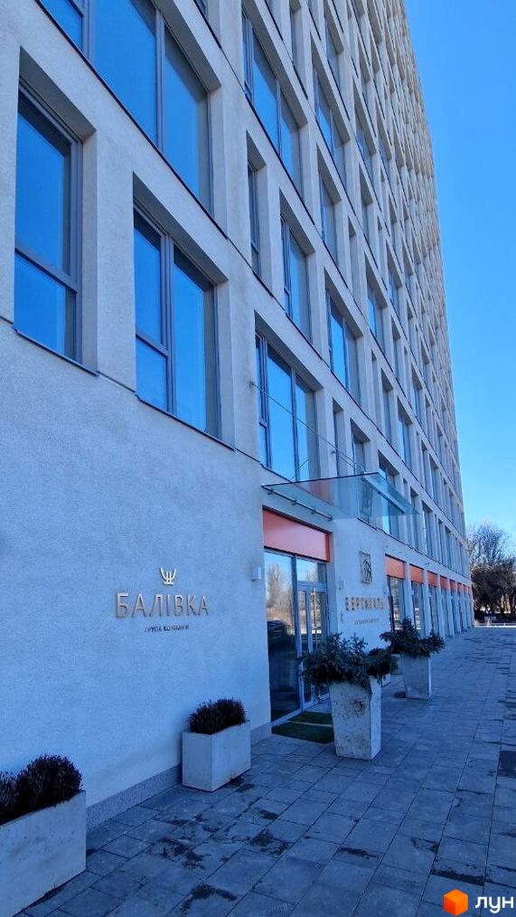 Фасад житлового комплексу Вертикаль з великими вікнами та вхідною групою, облаштована прибудинкова територія з плиткою та рослинами.