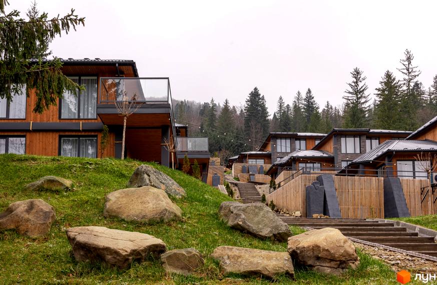 Котеджне містечко Агат Резорт & SPA: будинки з дерев'яними фасадами, темними дахами та великими вікнами. Територія з зеленими схилами, камінням та хвойним лісом.