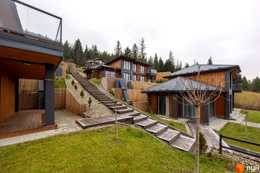 Котеджне містечко Агат Резорт & SPA: будинки з дерев'яними фасадами та темними дахами на схилі, з'єднані дерев'яними сходами та зеленими газонами.