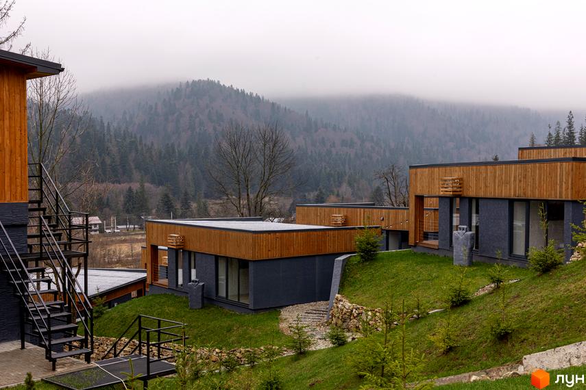 Котеджне містечко Агат Резорт & SPA: будинки з дерев'яним оздобленням та темними фасадами, оточені зеленими газонами, молодими деревами та горами.