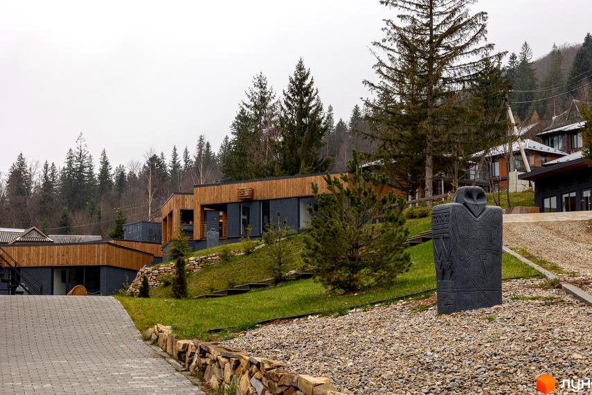 Котеджне містечко Агат Резорт & SPA з дерев'яними фасадами на схилі пагорба. Прибудинкова територія має зелені газони, кам'яні стіни та скульптуру.