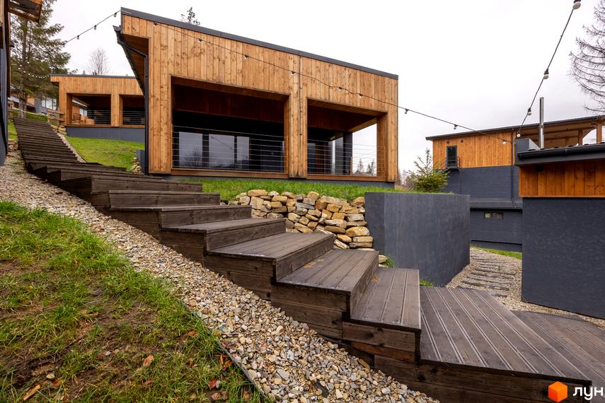 Дерев'яні будинки Котеджного містечка Агат Резорт & SPA з темними фасадами та великими вікнами, розташовані на схилі з дерев'яними сходами та газоном.