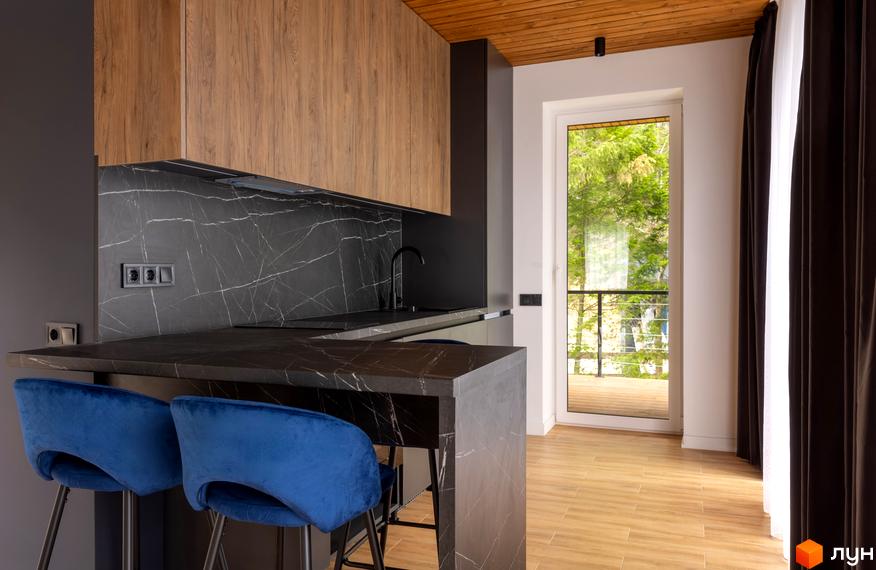 Кухня-вітальня з темною мармуровою стільницею та дерев'яними шафами. З вікна Котеджного містечка Агат Резорт & SPA видно зелені дерева.