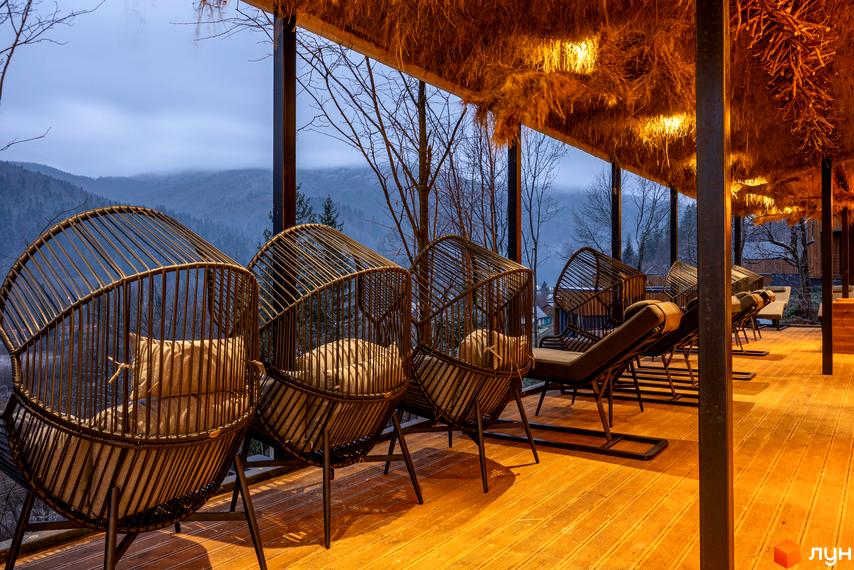 Котеджний комплекс КМ Agat Resort & SPA з плетеними кріслами та лежаками на терасі з видом на гори. Затишна зона відпочинку під солом'яним навісом.