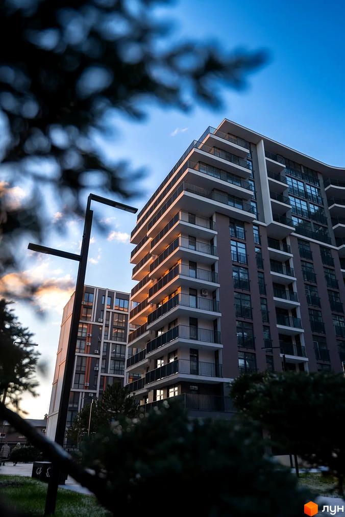 Житловий комплекс Crystal - сучасна новобудова з балконами та панорамними вікнами на фоні блакитного неба. Навколо зелені насадження та освітлення.