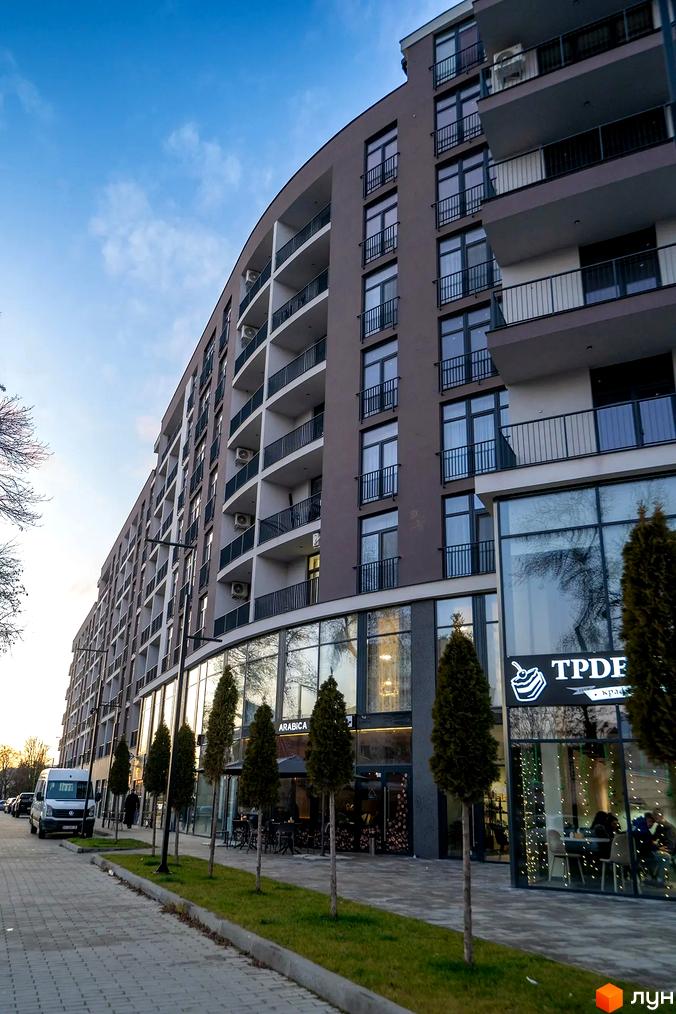 Багатоповерховий Житловий комплекс Crystal з вигнутим фасадом, балконами та великими вікнами на першому поверсі. Перед будинком облаштована пішохідна зона з деревами.