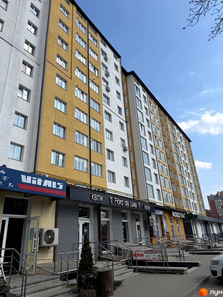 Фасад багатоповерхового будинку з жовто-білими секціями та комерційними приміщеннями на першому поверсі. Навколо будинку облаштована прибудинкова територія з тротуаром та зеленими насадженнями.