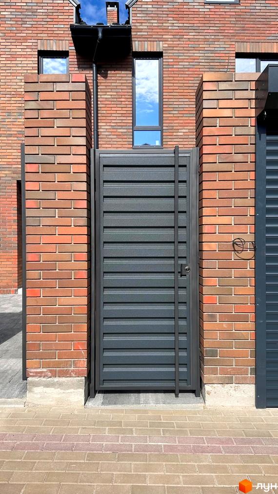 Фасад будинку з цегляним оздобленням та металевою брамою. Поруч розташована бруківка та зелена зона.