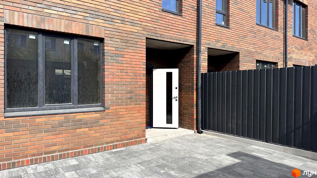 Фасад будинку з цегляним оздобленням, білими дверима та чорним парканом. Прибудинкова територія з бруківкою.