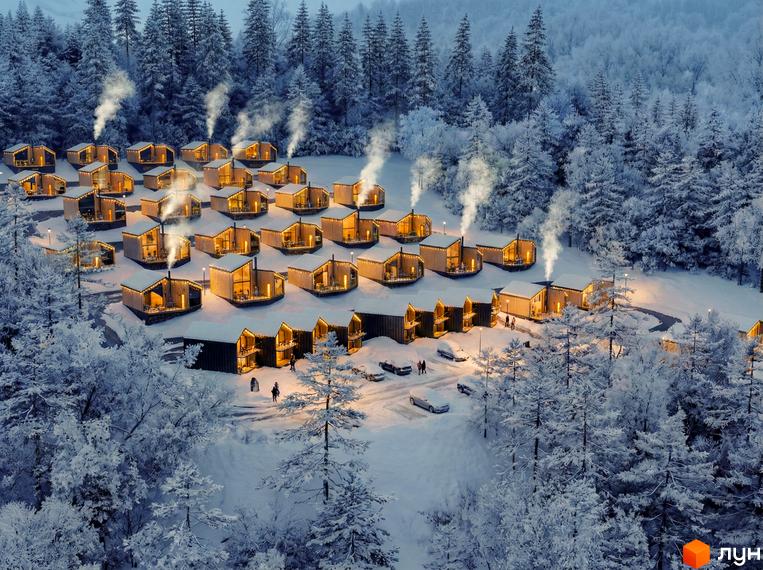 Котеджне містечко Теплий: дерев'яні будинки з панорамними вікнами та димом з димоходів на тлі зимового лісу. Територія комплексу вкрита снігом, освітлена ліхтарями та гірляндами.