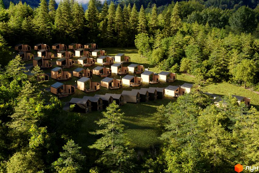 Котеджне містечко Теплий: дерев'яні будинки з темними дахами на зеленому схилі серед лісу. Навколо багато дерев, видно доріжки та освітлення.