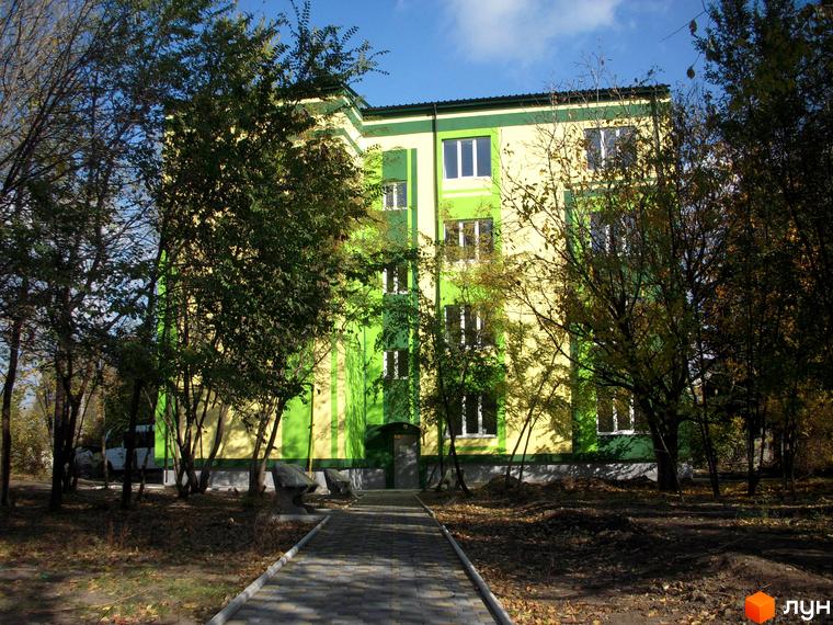 Зовнішній вигляд новобудови ЖК Усадьба з жовто-зеленим фасадом. Навколо будинку розташовані дерева та доріжка.