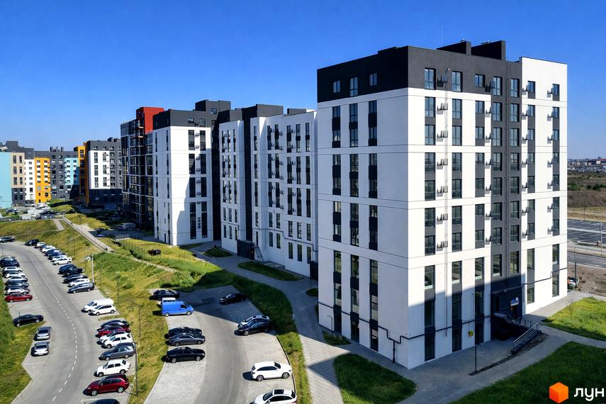 Багатоповерхові будинки Житлового комплексу На Щасливому з білими фасадами та темними вікнами, поруч парковки з автомобілями та зелені газони.