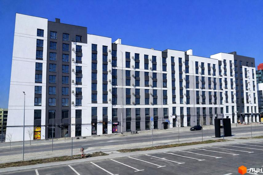 Житловий комплекс На Щасливому: багатоповерховий будинок з білим та темно-сірим фасадом, численними вікнами та кондиціонерами. Перед будинком дорога та паркінг.