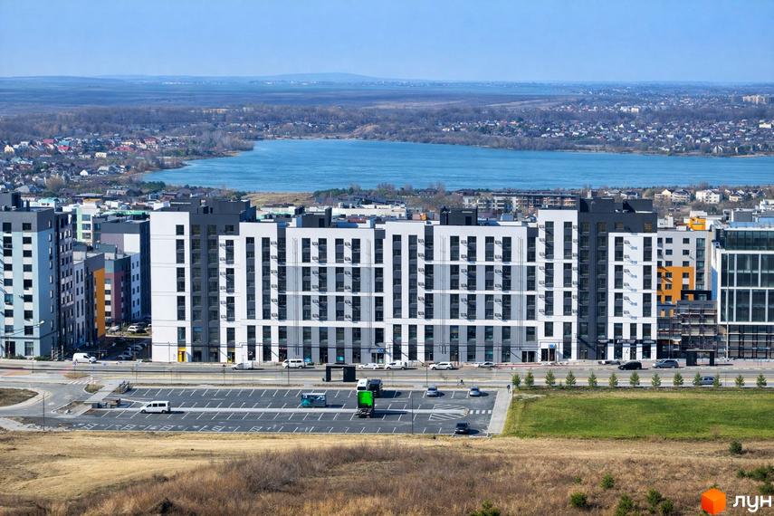 Житловий комплекс На Щасливому з багатоповерховими будинками білого, сірого та темного кольорів. Перед комплексом парковка, а на горизонті видно озеро.