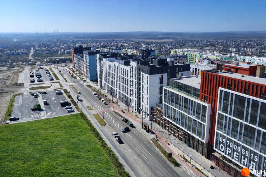 Житловий комплекс На Щасливому з багатоповерховими будинками білого, сірого, синього та помаранчевого кольорів. Поруч дорога, парковки та зелені насадження.