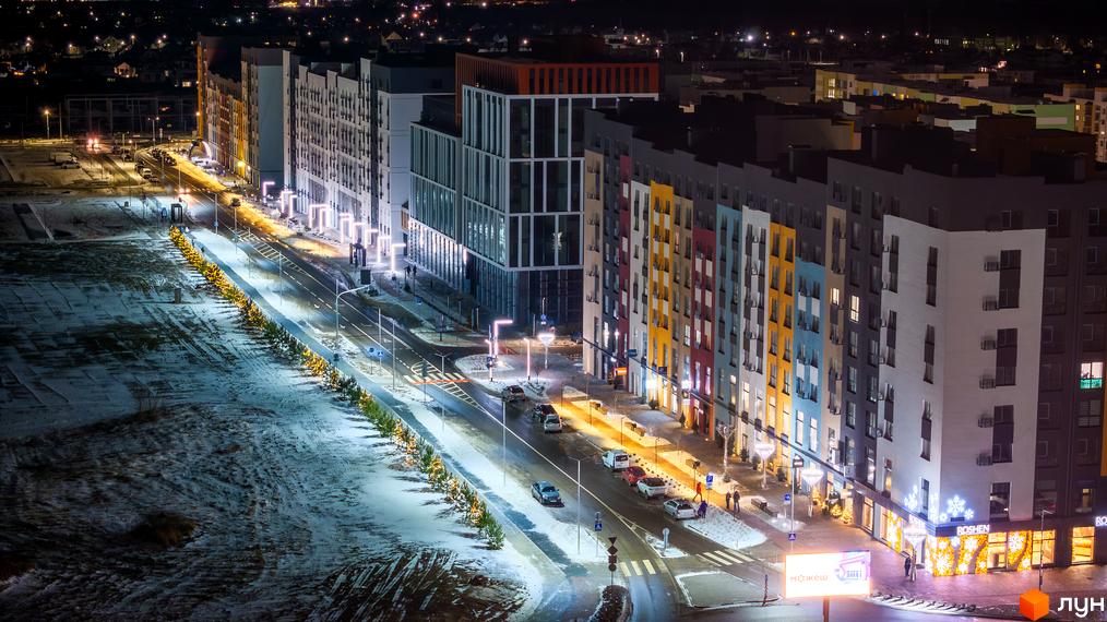 Житловий комплекс На Щасливому вночі, з фасадами різнокольорових будинків та освітленою вулицею. Перед будинками розташована зелена зона та дорога з автомобілями.
