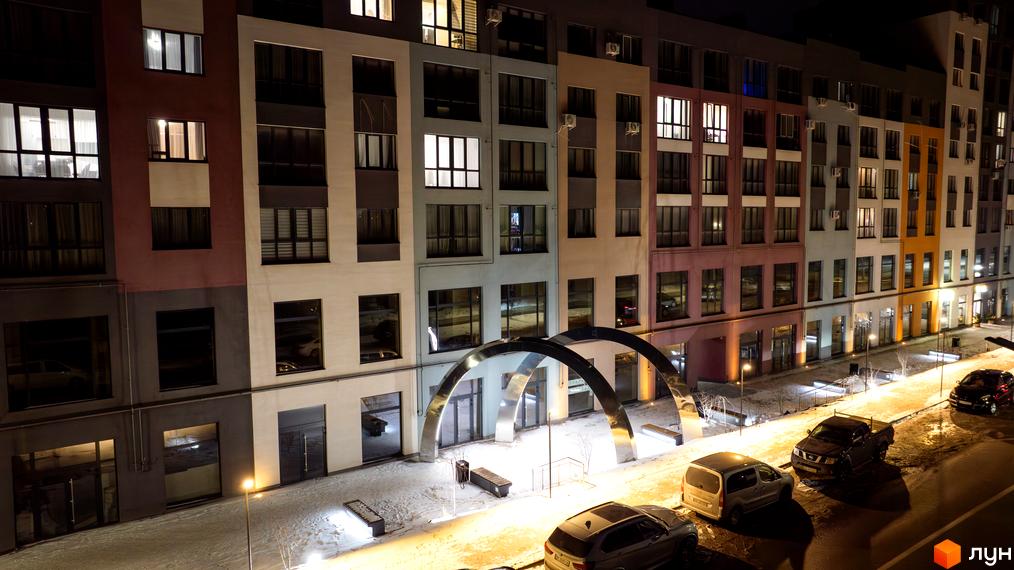 Житловий комплекс На Щасливому. Фасади будівель з підсвіченими вікнами та прибудинкова територія з арками та лавками вночі.