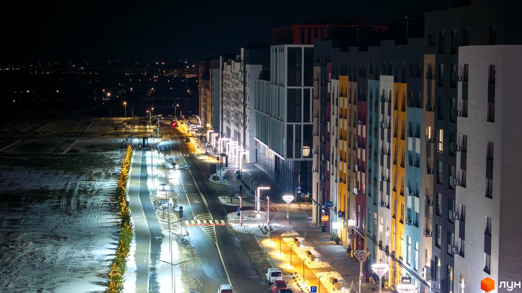 Житловий комплекс На Щасливому вночі з яскравим фасадом та освітленою вулицею. Поруч зелені насадження з новорічними гірляндами та снігове покриття.