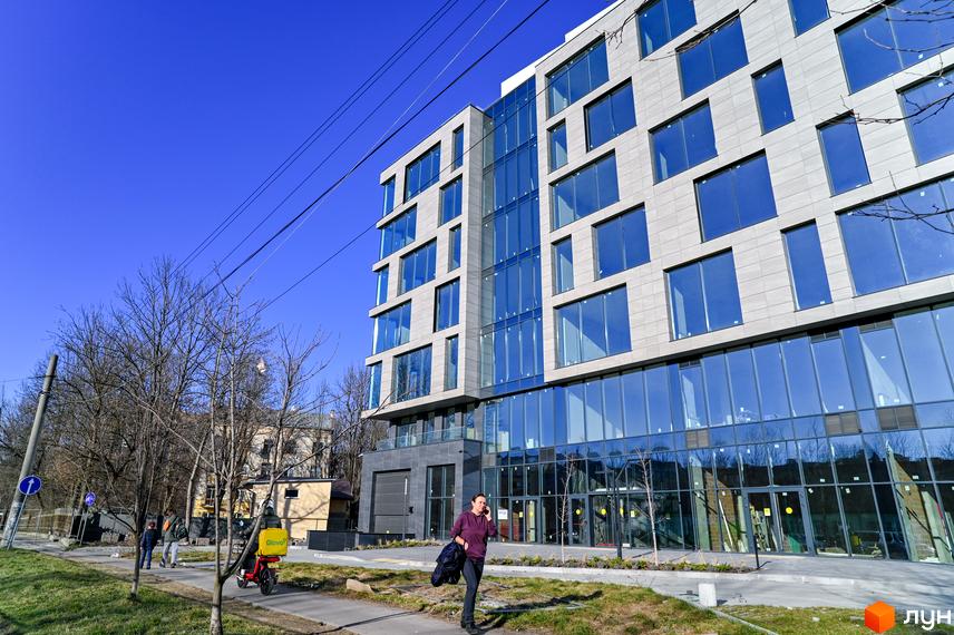 Будівельний проєкт Mill Town з фасадом з великих вікон та сірих панелей, поруч зелена зона та тротуар.