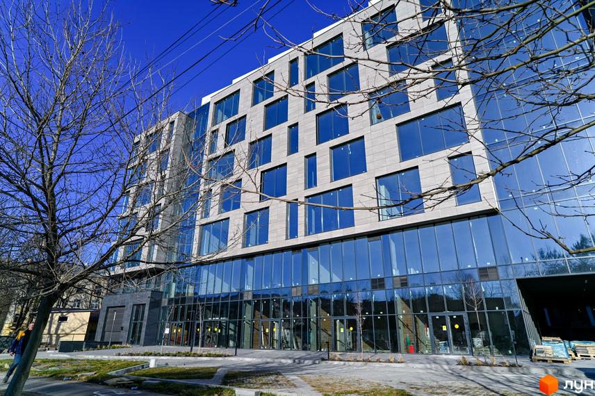 Фасад новобудови ЖК Mill Town з панорамними вікнами та сірою облицовкою, перед ним розташована прибудинкова територія з деревами.